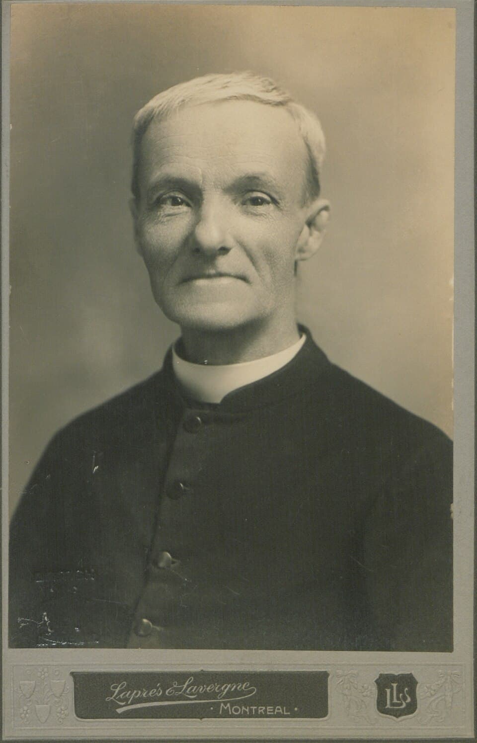 Studio photograph of Brother André Bessette as an older man with short white hair, smiling slightly, wearing a black buttoned cassock with a white collar, mounted on a card stamped Laprés & Lavergne, Montreal
