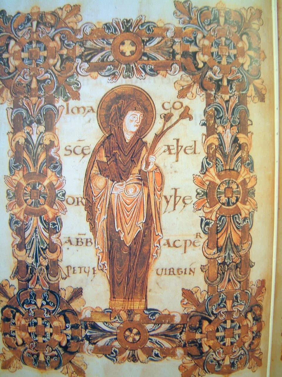 Manuscript painting of Saint Etheldreda standing in a brown veil and robes with a gold halo, holding a book and a flowering branch, inside a gold border of leaves and rosettes with her name lettered on either side