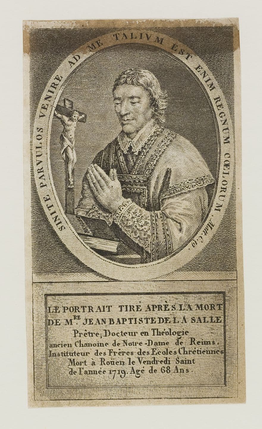 An engraved portrait of John Baptist de la Salle in a lace surplice, hands joined in prayer before a crucifix on a table, taken after his death in 1719