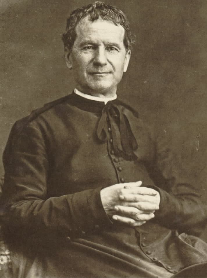 A photograph of Don Bosco in Turin in 1880, an elderly priest in a cassock and cloak, hands loosely clasped, looking slightly away from the camera