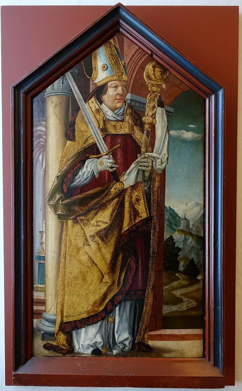 A painted wooden panel in a pointed frame showing a bishop in a white miter and gold cope, holding a sword across his body in one gloved hand and a gilded crozier in the other, standing beside a stone column with mountains and a castle behind him