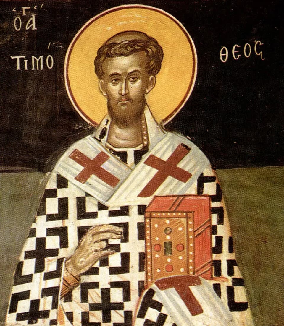 A Byzantine fresco of a bearded young bishop with a gold halo, wearing a white vestment patterned with black crosses and holding a jeweled book of the Gospels, his name written in Greek beside his head