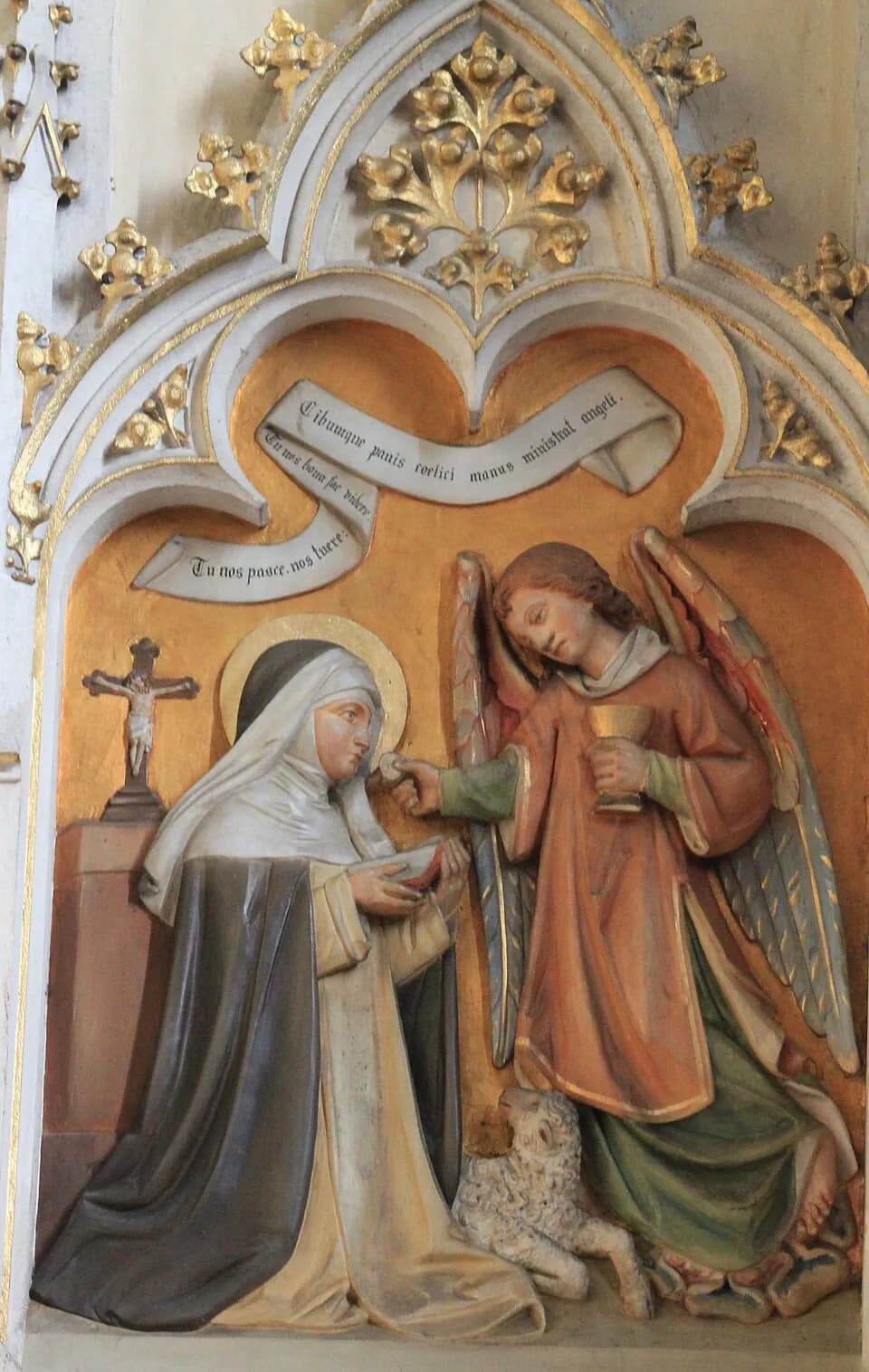 Painted and gilded relief of a kneeling Dominican nun with a halo receiving communion from a standing angel who holds a chalice, with a lamb at their feet, a crucifix behind her and a Latin scroll above, set in a Gothic arch