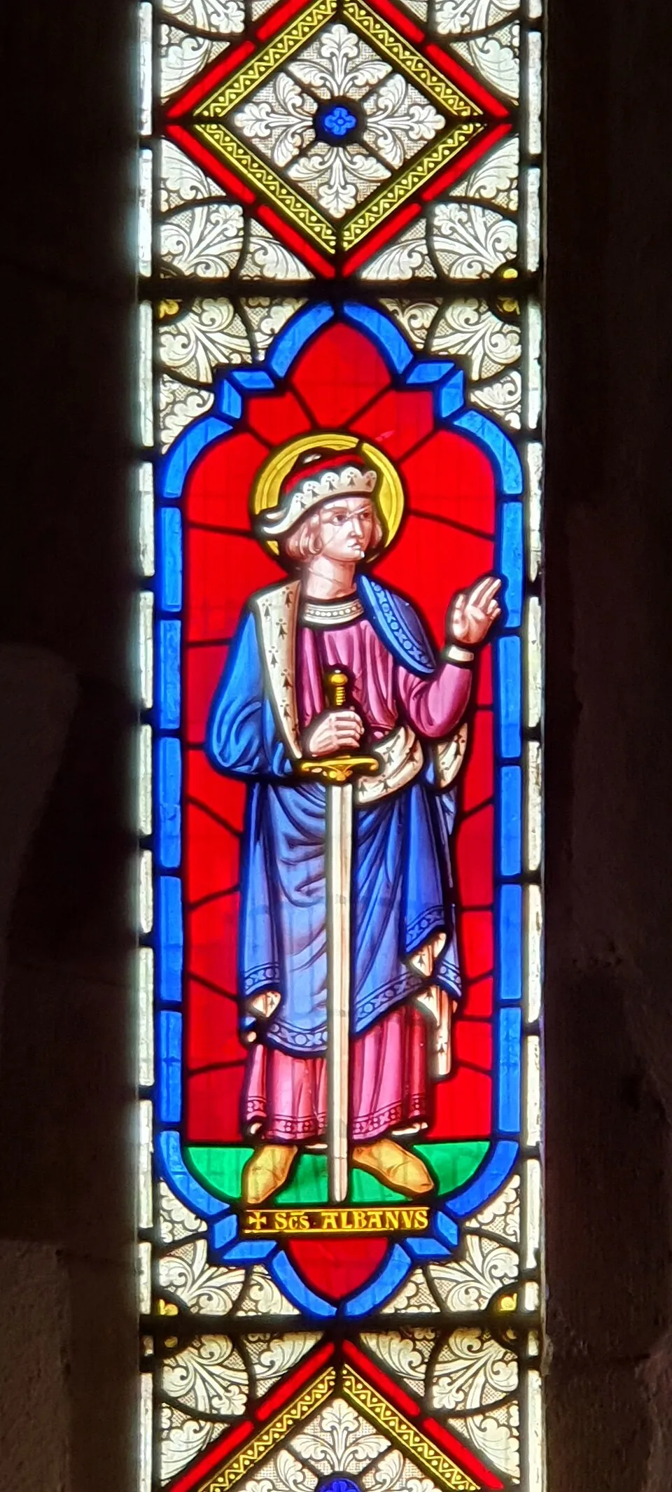 A Victorian stained-glass figure of Saint Alban as a young man with a halo and a fur-trimmed cap, in a blue cloak over a pink tunic, holding the hilt of a long sword point-down and raising his right hand, on a red ground, above a label reading Scs Albanus