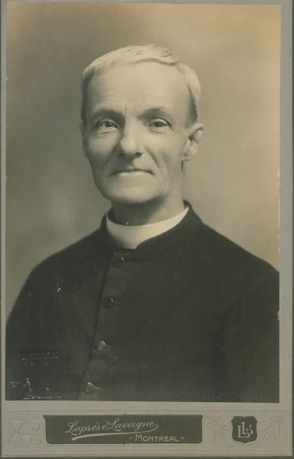 Fotografía de estudio del hermano André Bessette ya mayor, de cabello blanco y corto, con una leve sonrisa, vestido con una sotana negra abotonada y cuello blanco, montada en un cartón con el sello de Laprés & Lavergne, Montreal