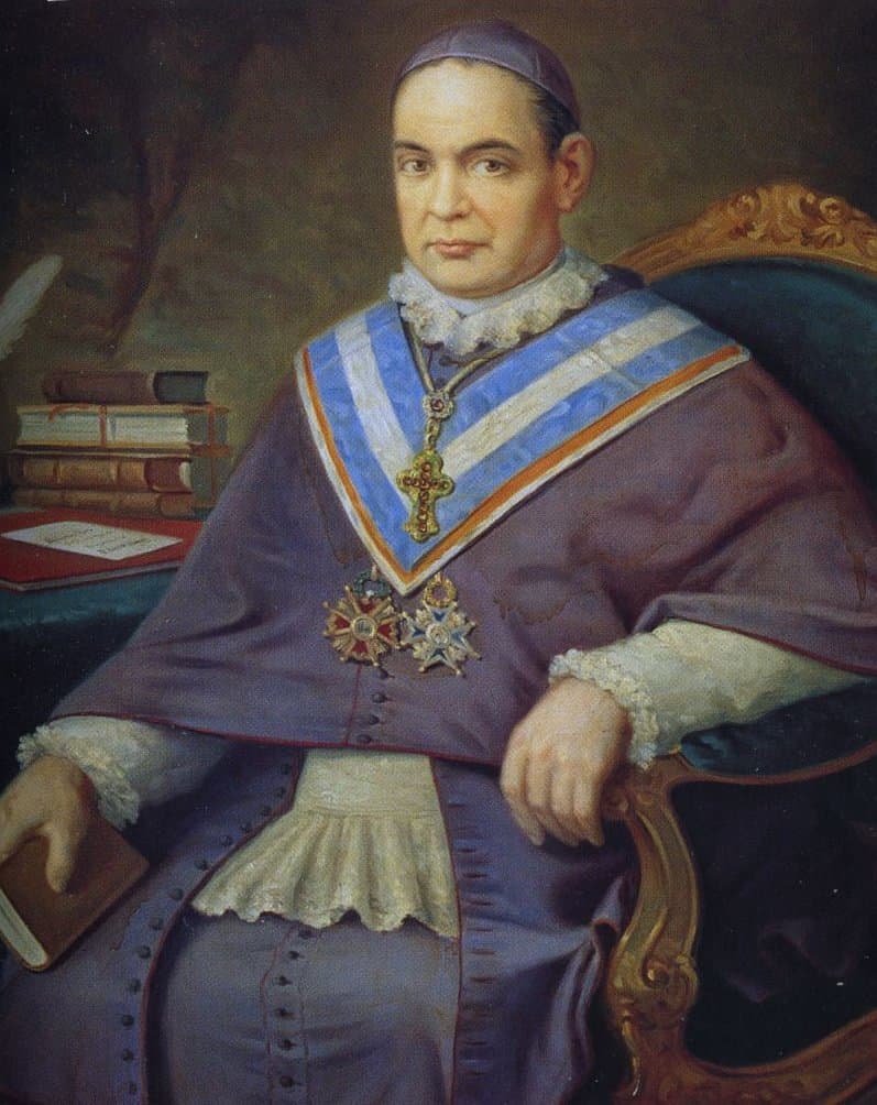 Anthony Mary Claret seated in a gilded armchair in purple archbishop's dress, wearing a pectoral cross, a blue and white sash and two medals, with books on a table behind him