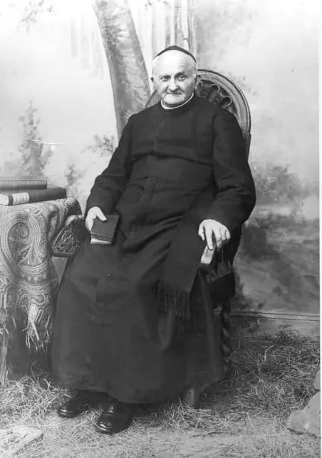A black-and-white studio photograph of Arnold Janssen as an old man, in a black cassock, sash and skullcap, seated in a carved chair with a small book in one hand, before a painted backdrop of trees
