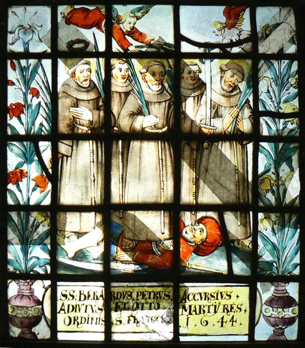 Stained-glass window of five tonsured friars in brown habits holding martyrs' palms and swords, standing over a fallen king in a red turban, with angels bringing crowns from above, lilies at the sides and a Latin inscription naming Berard, Peter, Accursius, Adjutus and Otto