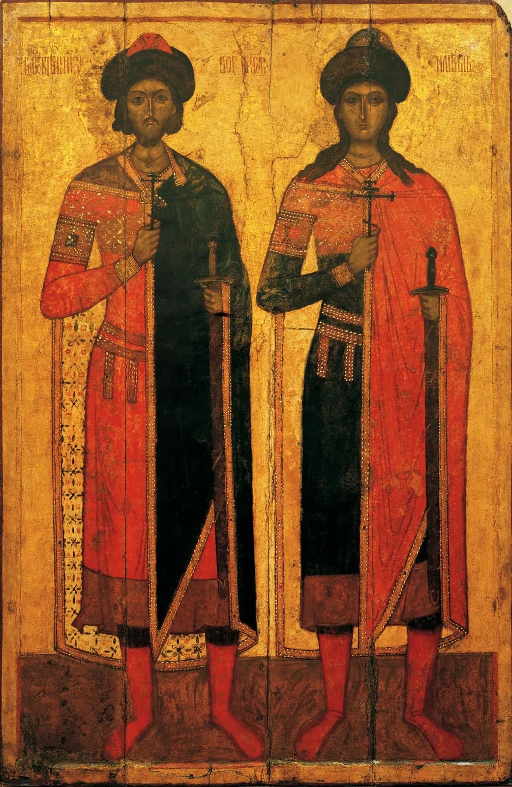 Medieval Russian icon of two young princes standing side by side on a gold ground, both in fur-trimmed caps and long red cloaks, each holding a small cross in his right hand and a sheathed sword in his left, the older one bearded and the younger beardless, with faded inscriptions above their heads