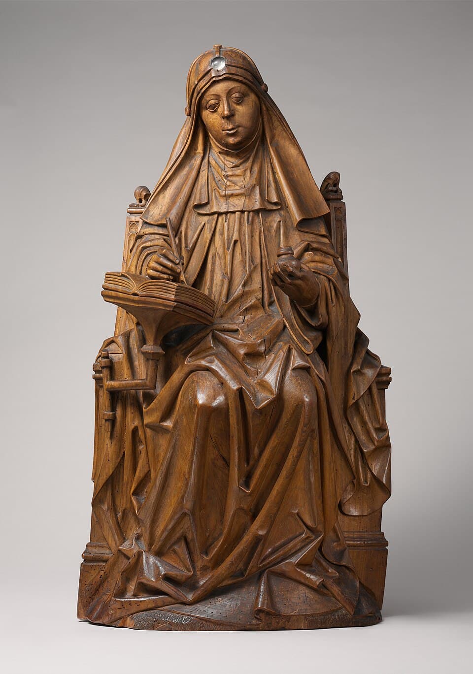 A carved wooden figure of Saint Bridget of Sweden seated in a high-backed chair in a widow's veil and wimple, writing with a quill in an open book on a stand