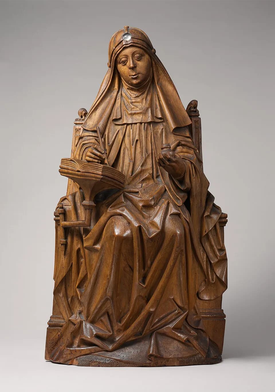Figura de madera tallada de Santa Brígida de Suecia, sentada en una silla de respaldo alto, con velo y toca de viuda, escribiendo con una pluma en un libro abierto sobre un atril