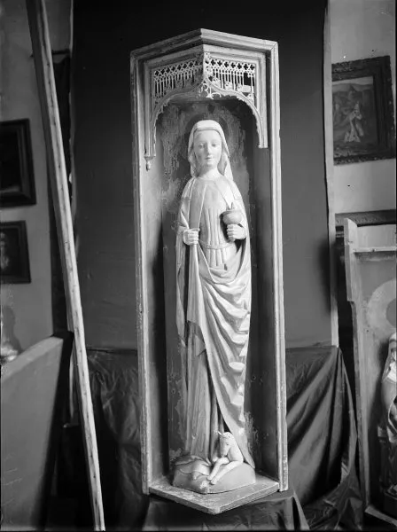Black-and-white photograph of a carved wooden statue of a woman in a veil and long mantle standing in a tall niche under a pierced Gothic canopy, holding a small vessel in one hand, with a small deer lying at her feet