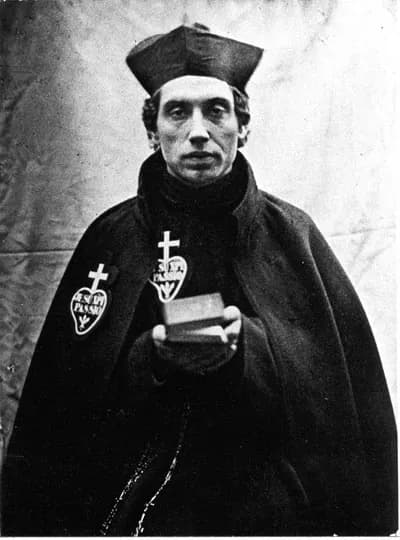 A black-and-white photograph of Father Charles Houben as a young Passionist priest, in a biretta and dark cloak bearing the heart-shaped Passionist badge, holding a small book in front of him