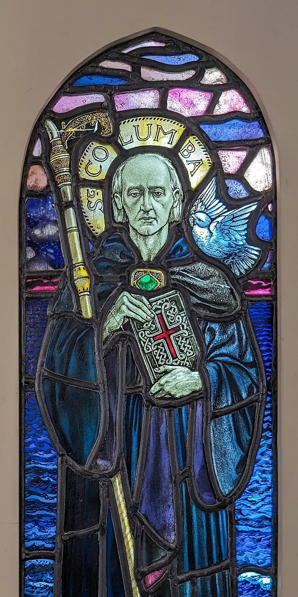 Stained glass window of Saint Columba as a gray-haired abbot in a dark blue cowl, holding a crozier and a jeweled book, with a white dove at his shoulder, his name in his halo, and waves behind him