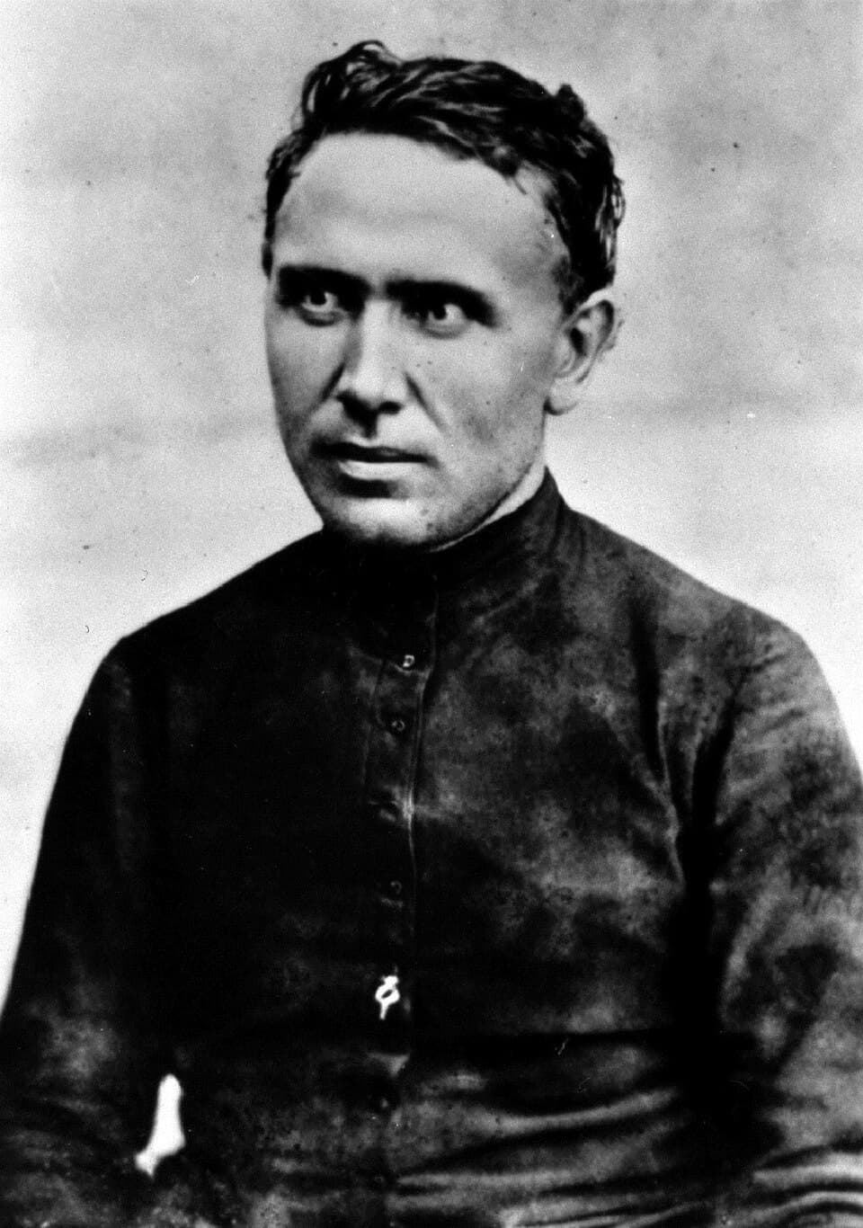 A black-and-white photograph of Father Damien as a young priest, dark-haired, in a buttoned black cassock, looking slightly to one side