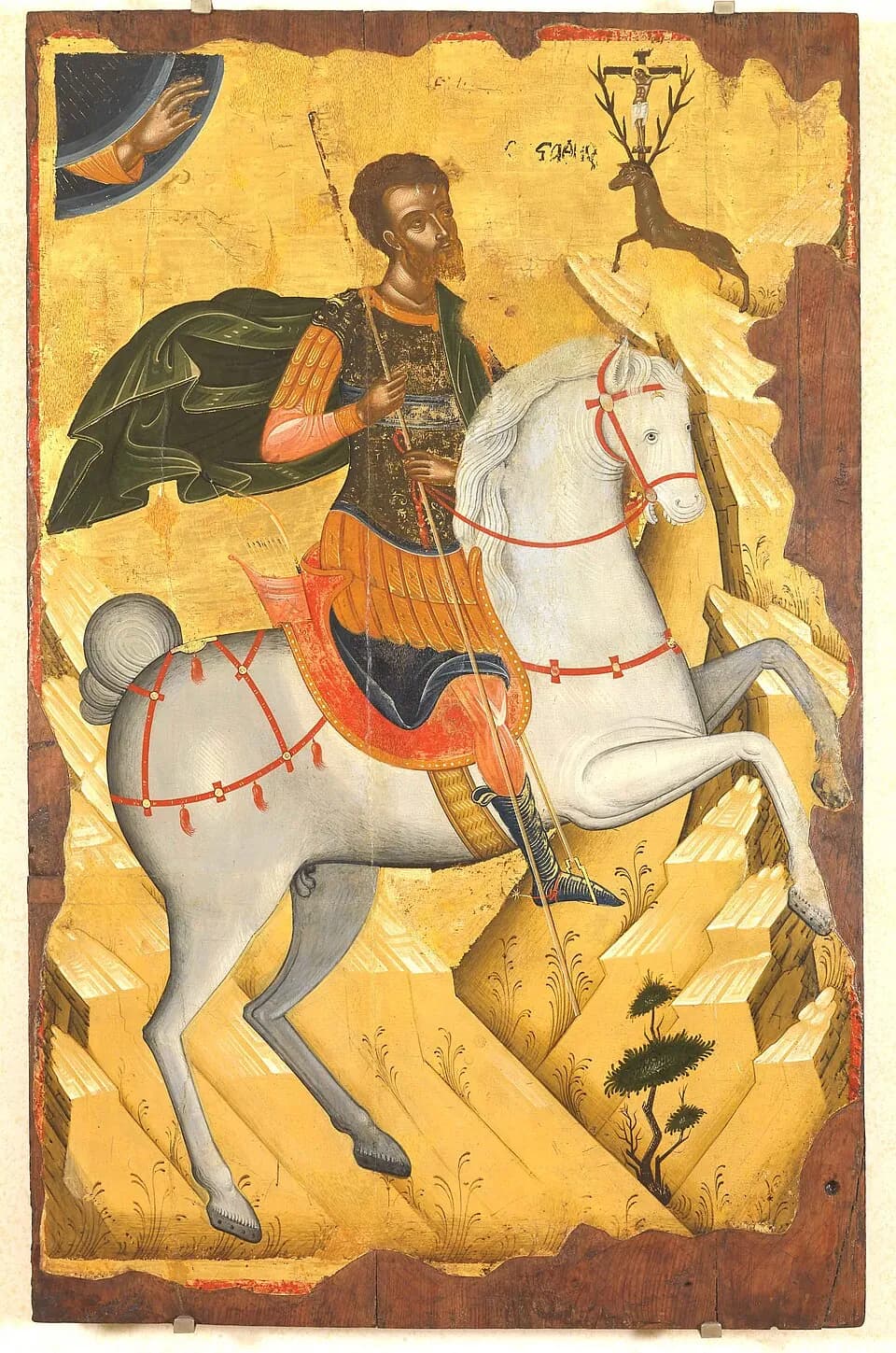 A Greek icon on a gold ground: a bearded soldier in Roman armor and a green cloak rides a rearing white horse and looks up at a stag on a rocky ledge with the crucified Christ standing between its antlers, while the hand of God blesses from the upper corner