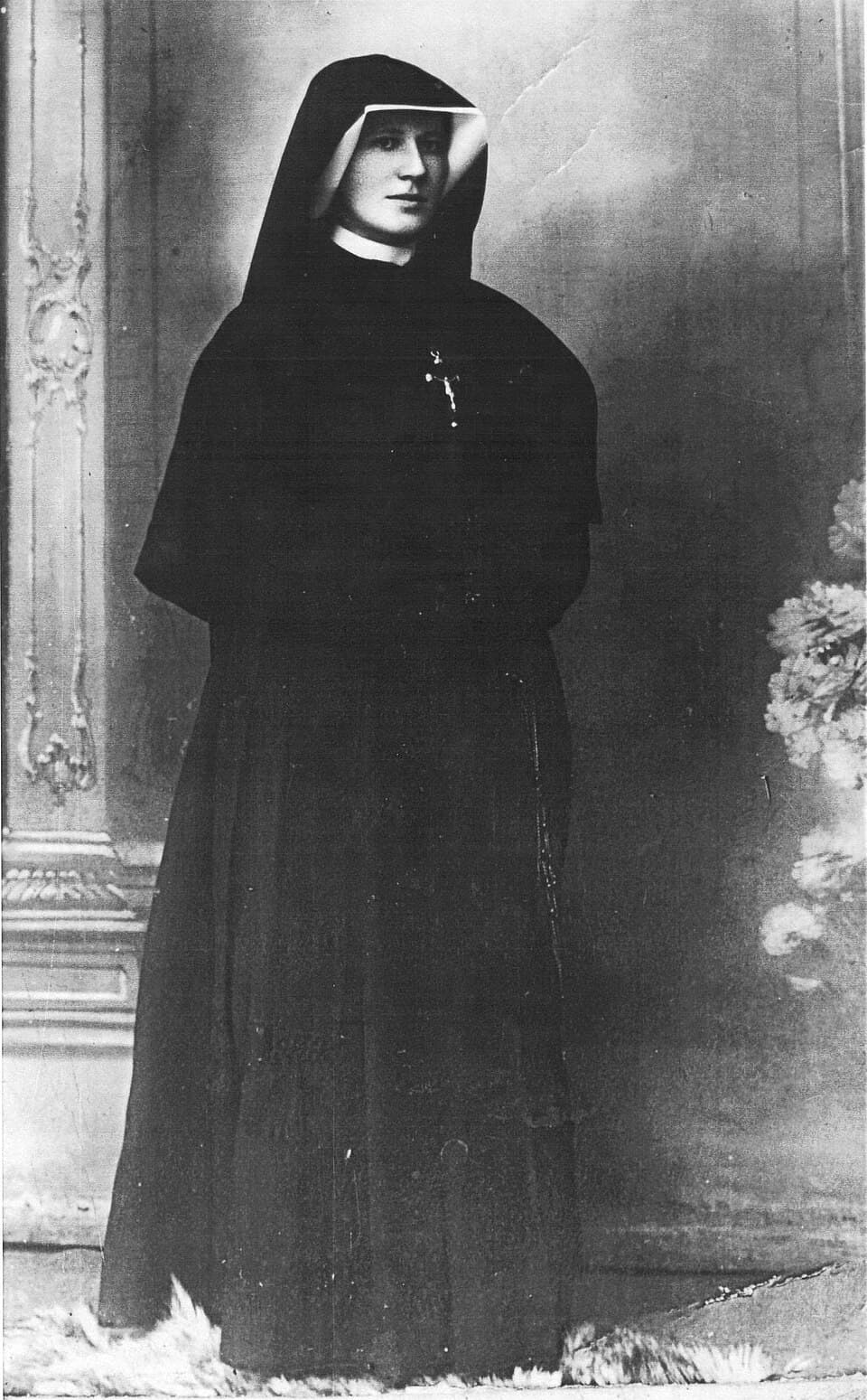 Black-and-white studio photograph of Sister Faustina standing full length in a dark habit and veil, a crucifix on her breast, against a painted backdrop