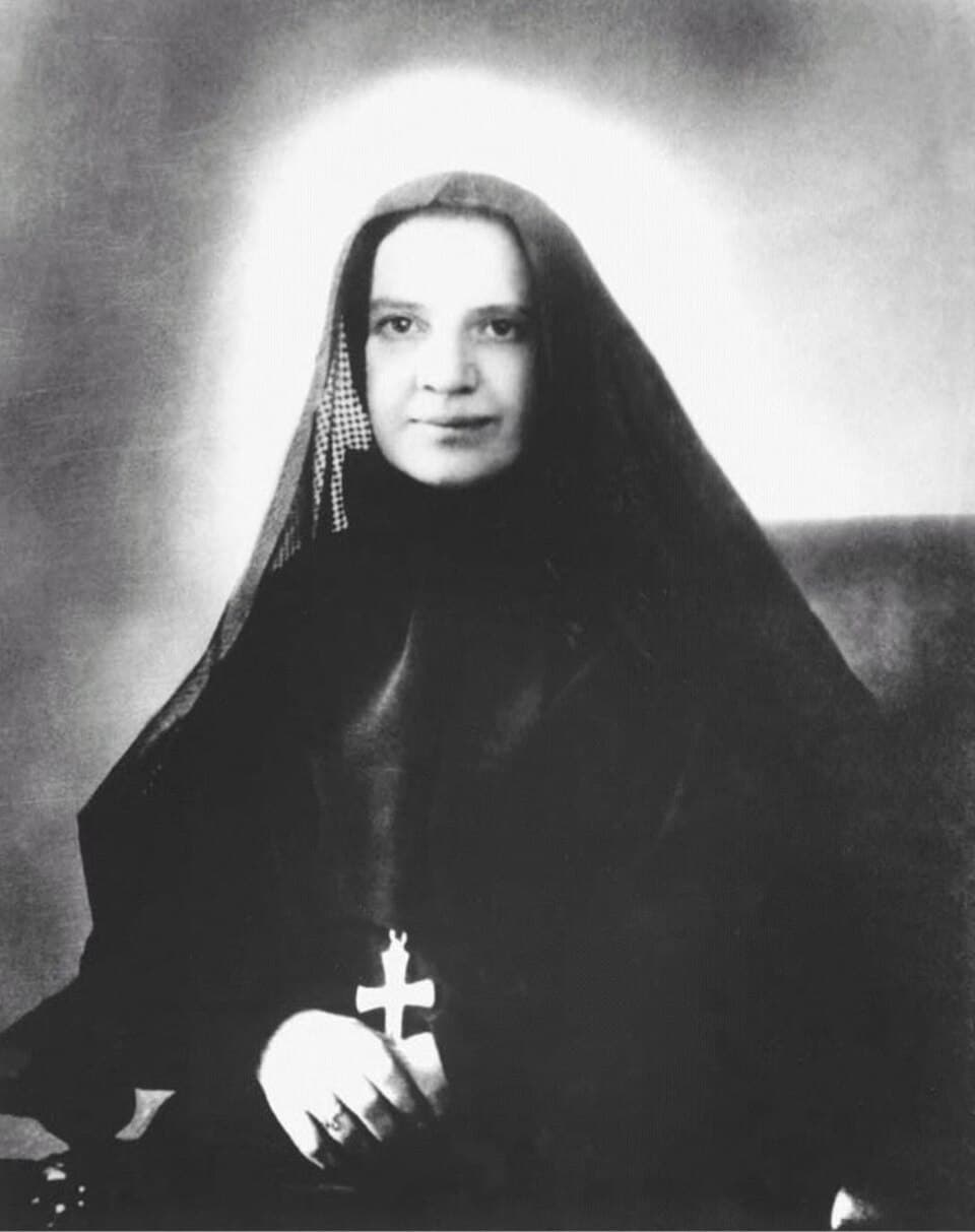 Black and white photograph of Mother Cabrini, seated, in a dark veil and habit with a cross at her breast, a pale halo-like glow behind her head