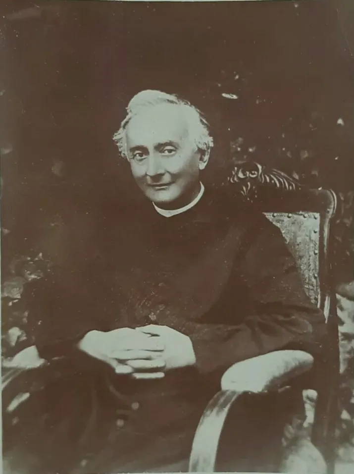 A sepia photograph of Francesco Spinelli as an older priest with white hair, seated in a carved armchair in a black cassock, his hands folded in his lap