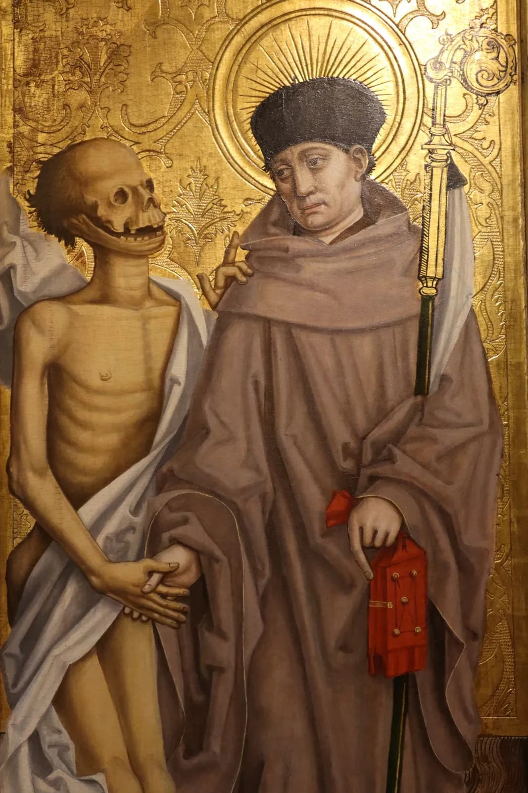 Late fifteenth-century panel painting on a tooled gold background of a haloed monk in a brown habit and dark cap, holding an abbot's staff and a red book, who leads by the hand a gaunt, half-decayed corpse wrapped in a white shroud