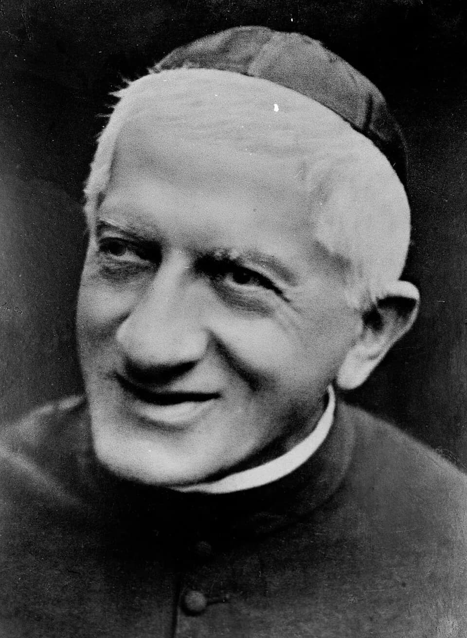 Black-and-white photograph of Giuseppe Allamano as an elderly priest in a cassock and skullcap, white-haired and smiling, looking to one side