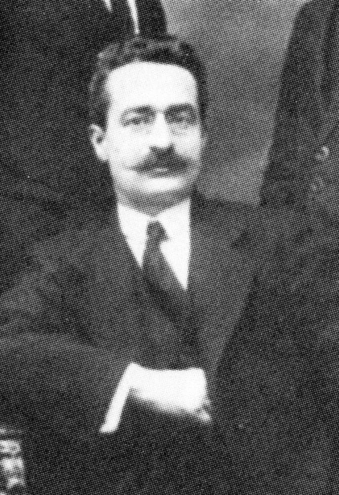A cropped detail of a black-and-white studio group photograph: Giuseppe Moscati, seated, in a dark suit and tie, with wire-rimmed glasses, a full dark mustache and dark hair brushed back, his hands resting together in his lap