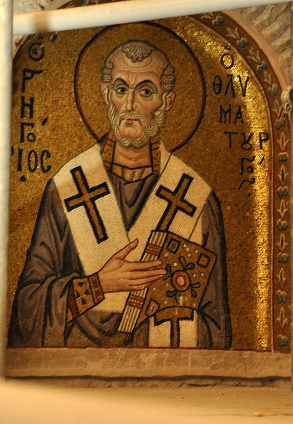 A Byzantine gold-ground mosaic of a gray-bearded bishop in a white stole marked with black crosses, holding a jeweled book, with his name in Greek letters on either side of his halo