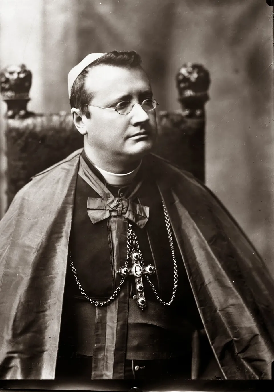 A black-and-white studio photograph of Guido Maria Conforti as a young archbishop, wearing round wire spectacles, a zucchetto, a silk mozzetta and a jeweled pectoral cross on a chain, seated before a carved chair