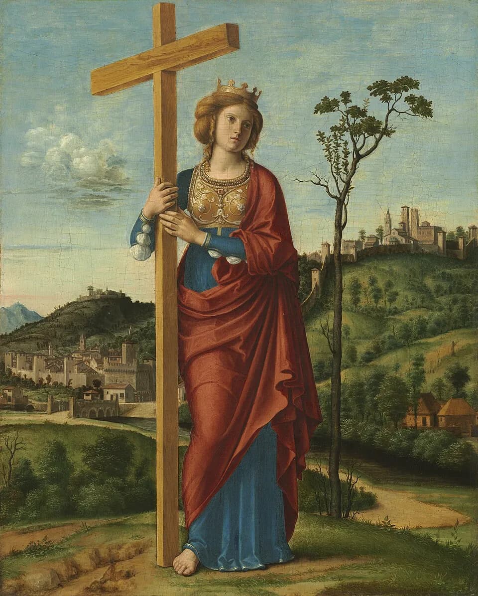 A crowned young woman in a red cloak and blue gown stands barefoot in a hilly landscape, holding a tall wooden cross upright beside her, with walled towns on the hills behind