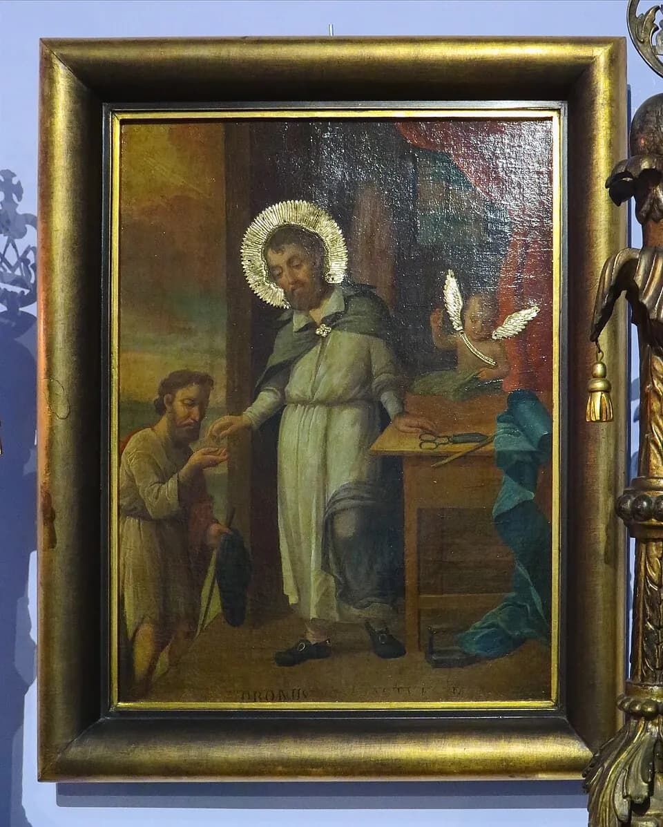 A haloed bearded man in a long pale robe drops a coin into the hand of a kneeling poor man, beside a table with tailor's shears, while a small winged angel looks on; the painting hangs in a gilded frame