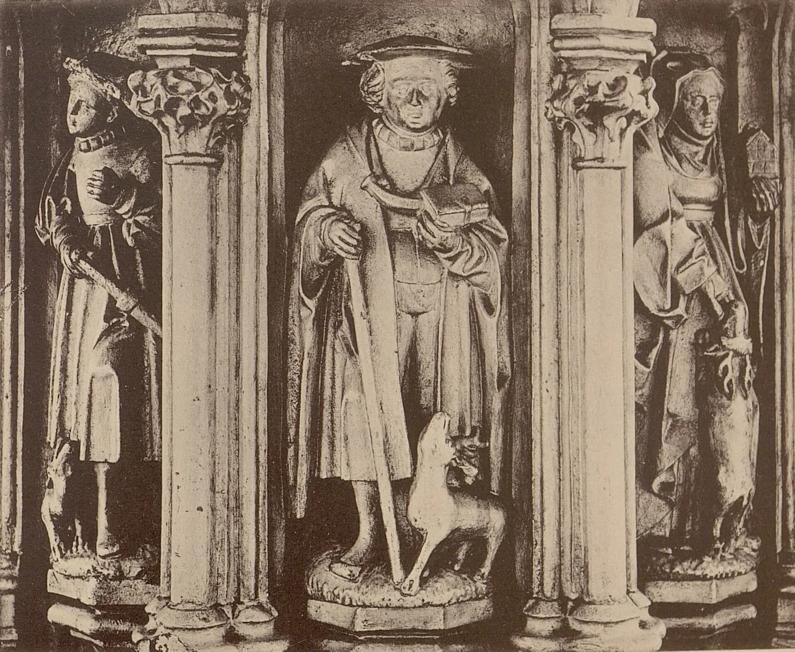 Antigua fotografía en sepia de tres santos tallados en piedra, de pie en hornacinas góticas entre columnas esbeltas; a la derecha una mujer con velo sostiene la maqueta de una iglesia mientras un ciervo se alza a su lado, en el centro un hombre con sombrero de ala ancha, bastón y libro tiene un ciervo a sus pies, y a la izquierda un joven con gorro sostiene una espada