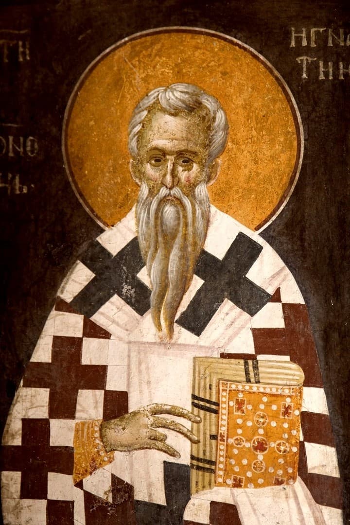 A Byzantine fresco of Saint Ignatius of Antioch as a white-bearded bishop in a cross-patterned omophorion, holding a jeweled gospel book, with his name lettered in Greek beside the halo