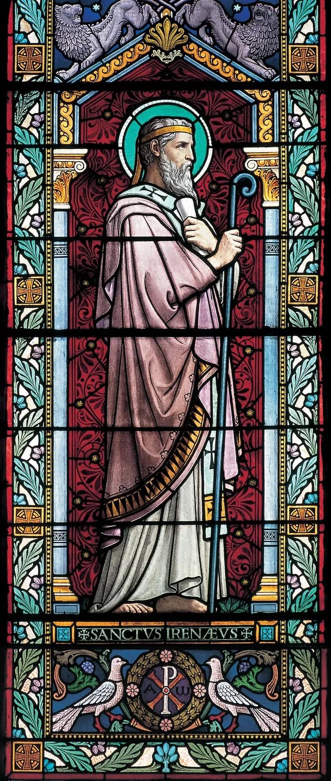 Una vidriera de Ireneo como obispo de barba gris con manto rosa, nimbo y una banda dorada en torno a la cabeza, que sostiene un rollo y un báculo, de pie sobre un fondo rojo con motivos y con la inscripción "Sanctus Irenaeus" a sus pies, y debajo un crismón entre dos palomas