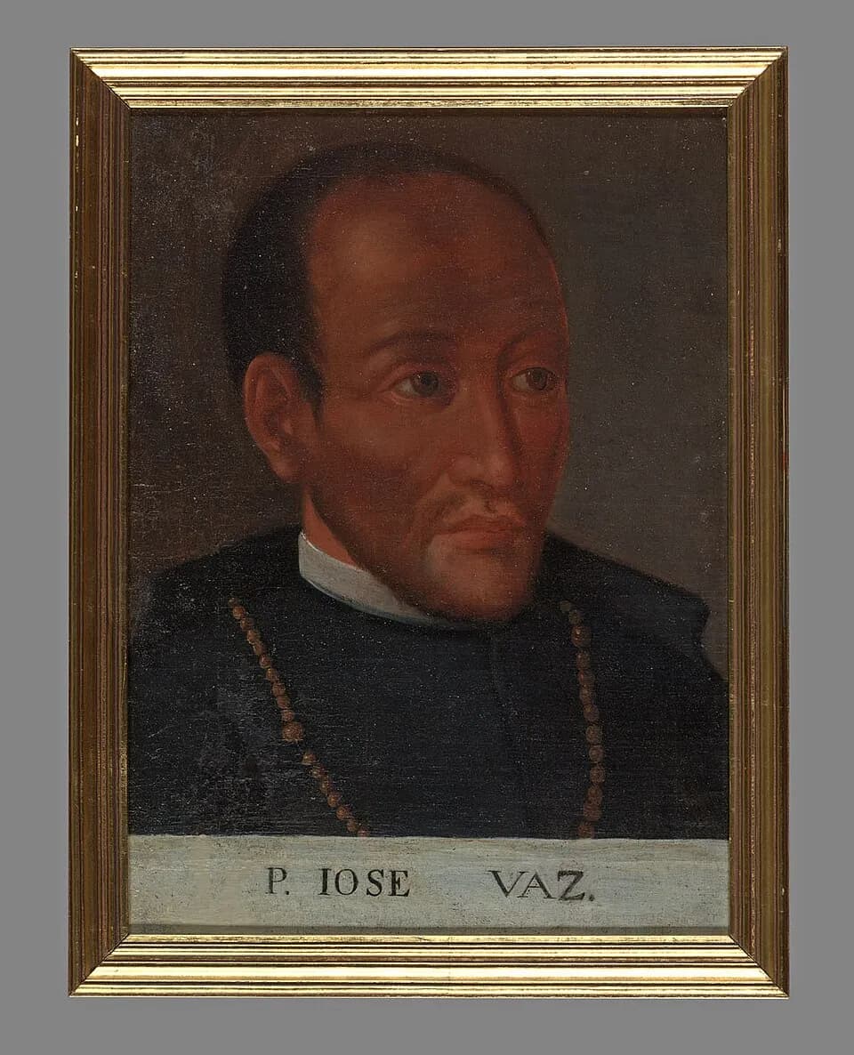 Framed oil portrait, head and shoulders, of a dark-skinned priest with a receding hairline and a thin mustache, in a black cassock with a white collar and a string of rosary beads around his neck, above a painted label reading P. IOSE VAZ