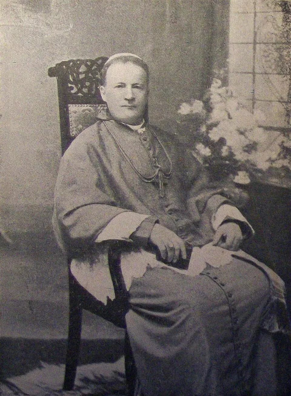 A black-and-white photograph of Archbishop Bilczewski seated in a carved wooden chair, in a bishop's choir dress with a pectoral cross on a chain and a skullcap, his hands resting on a book, with flowers beside him
