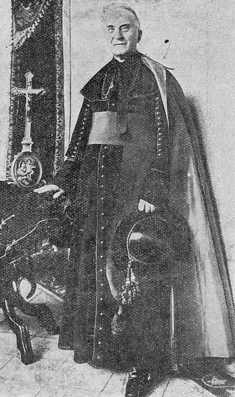 A black-and-white halftone photograph of Bishop Pelczar as an old man, standing full length in a bishop's cassock, sash and cape, one hand resting on a table beside a crucifix, the other holding a hat