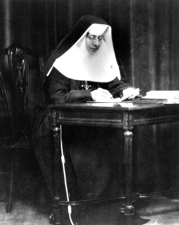 Katharine Drexel in the habit of her congregation, wearing glasses and writing at a carved wooden table