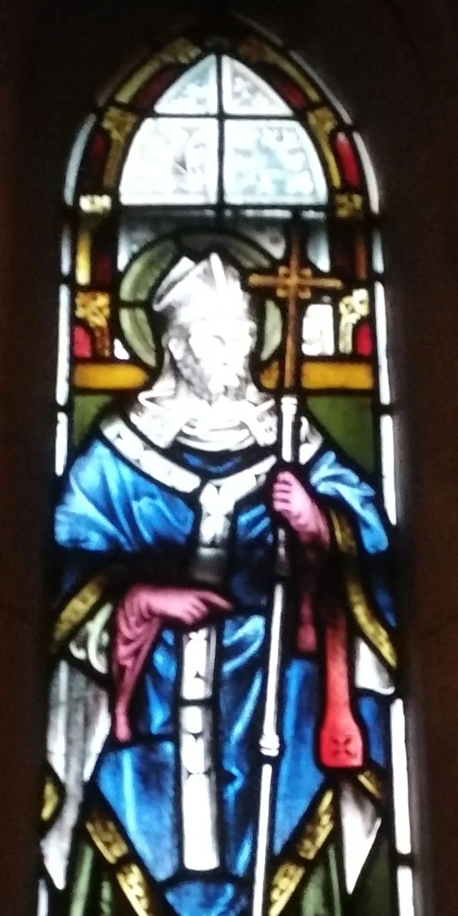 A stained-glass window of Saint Laurence O'Toole as a bearded archbishop with a halo and white miter, in blue vestments, holding a tall processional cross