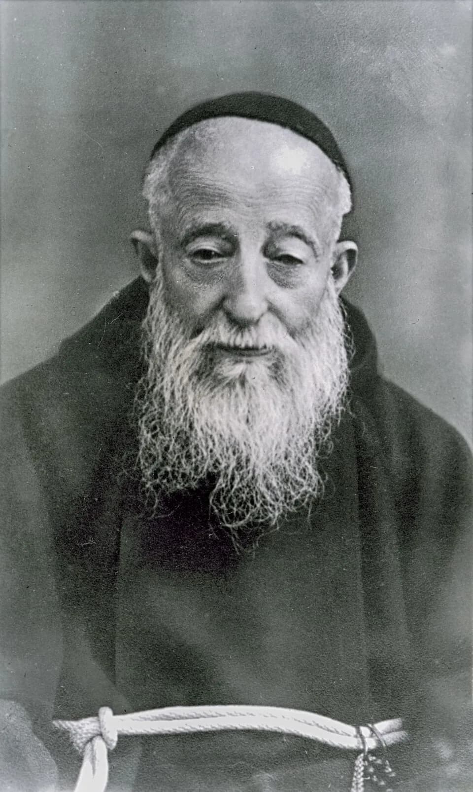 A black-and-white photograph of Leopold Mandić as an old man, in a Capuchin habit and skullcap, with a long white beard and a white cord at his waist, looking down