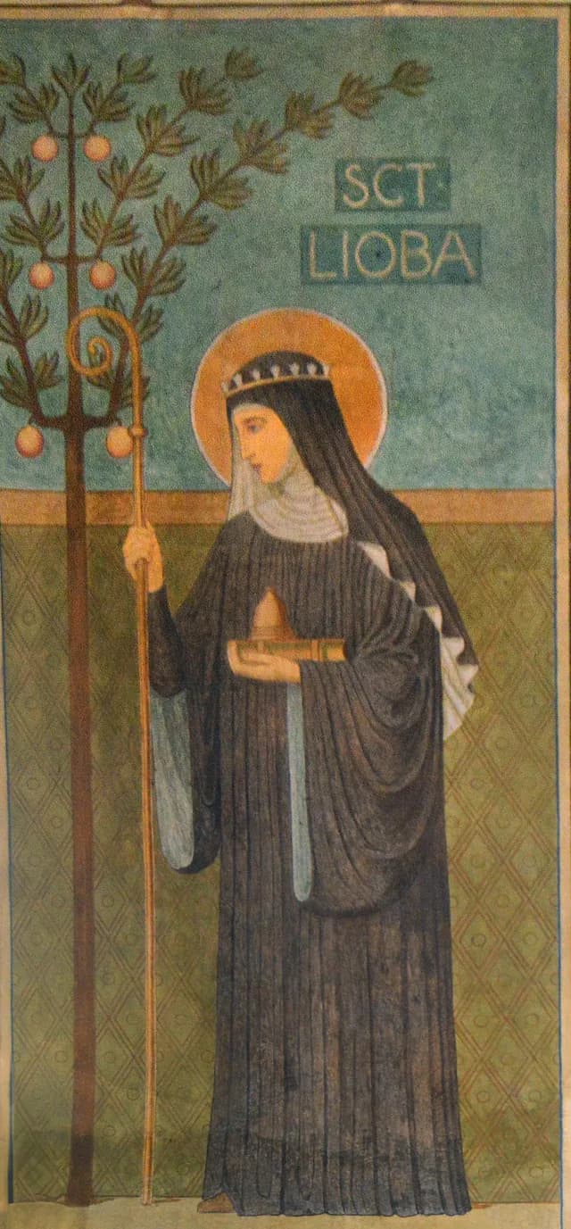 A painted panel of Saint Lioba as a Benedictine abbess in a black habit and gold halo, holding a crosier and a closed book with a small golden object on it, beside a fruit tree, with the words SCT LIOBA above her