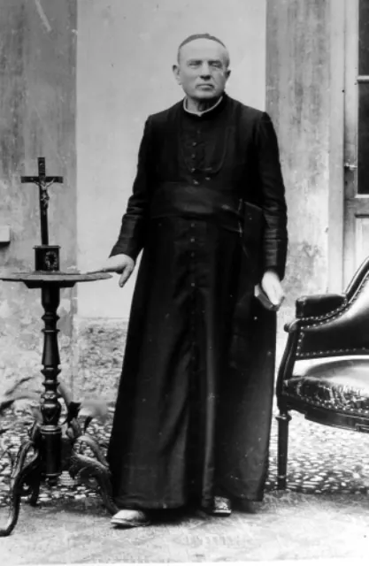 A black-and-white photograph of Don Luigi Guanella as an old man, standing full length in a black cassock in a courtyard, one hand resting on a small table with a crucifix and the other holding a book