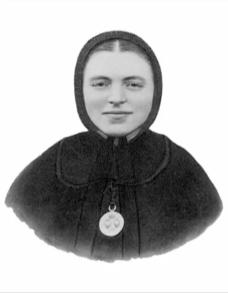 A black-and-white photograph of a young sister in a black veil and cape, smiling slightly, with a round medal of two hearts hanging at her chest