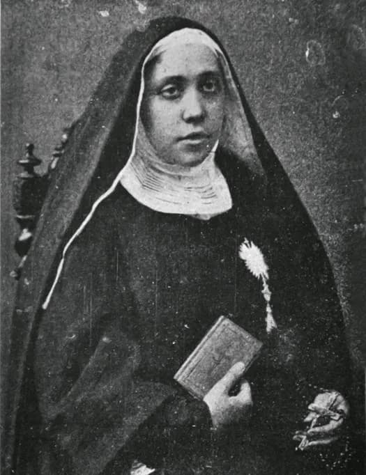 A black-and-white photograph of Maria Cristina Brando seated, in a dark habit and veil with a white wimple, a monstrance emblem pinned to her breast, holding a small book