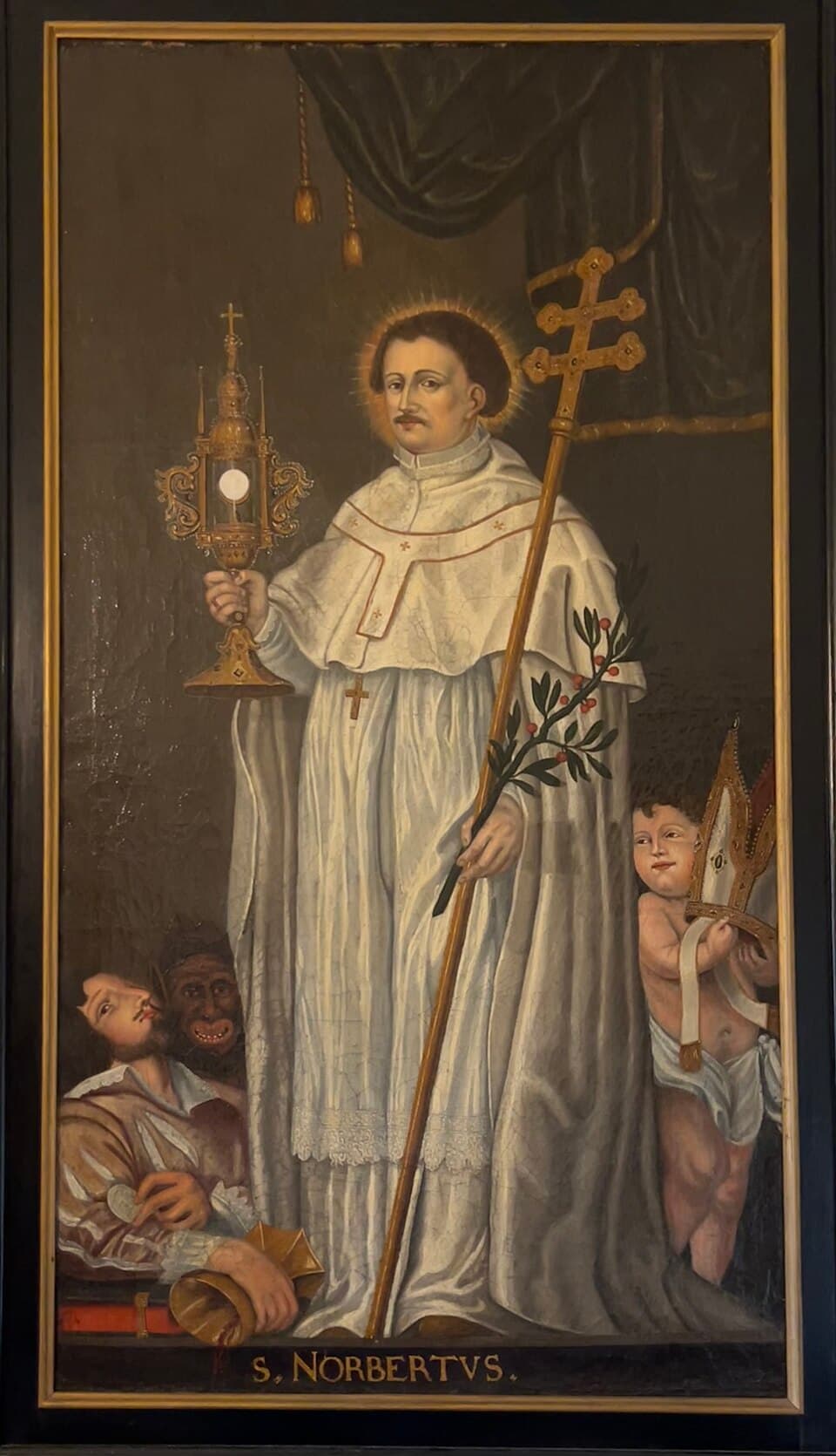A church painting labeled S. Norbertus, showing him in the white habit of a Premonstratensian canon holding a gold monstrance in one hand and an archbishop's cross-staff with an olive branch in the other, a defeated heretic at his feet and a cherub holding a mitre beside him