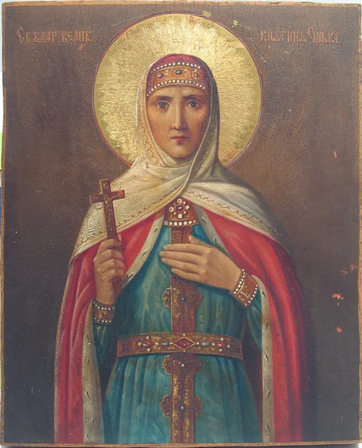 Russian icon of a crowned woman with a gold halo and a white veil over a jeweled headdress, wearing a red cloak over a blue-green gown, holding a small cross in her right hand and resting her left hand on her chest, against a dark brown background with an inscription in the upper corners