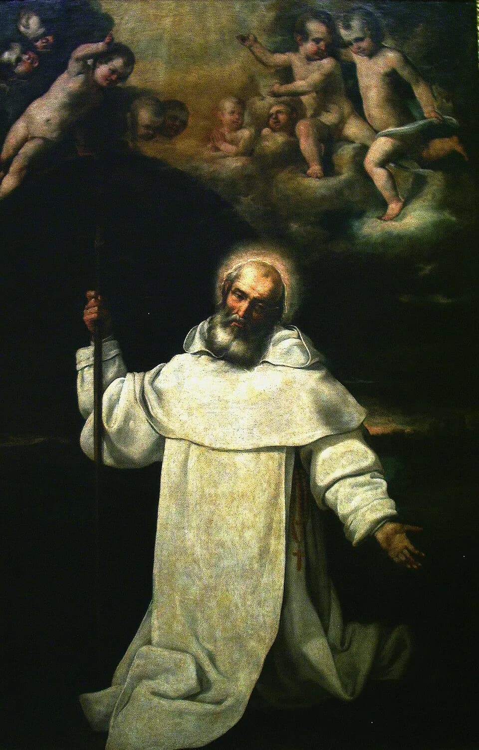 A gray-bearded friar with a halo, in a white habit and scapular, kneels holding a tall staff with his other hand held open, while cherubs gather in the clouds above him