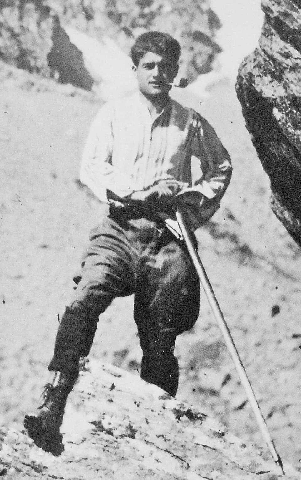 A black-and-white photograph of Pier Giorgio Frassati on a rocky mountainside, in a white shirt and climbing breeches, a pipe in his mouth and an ice axe planted beside him