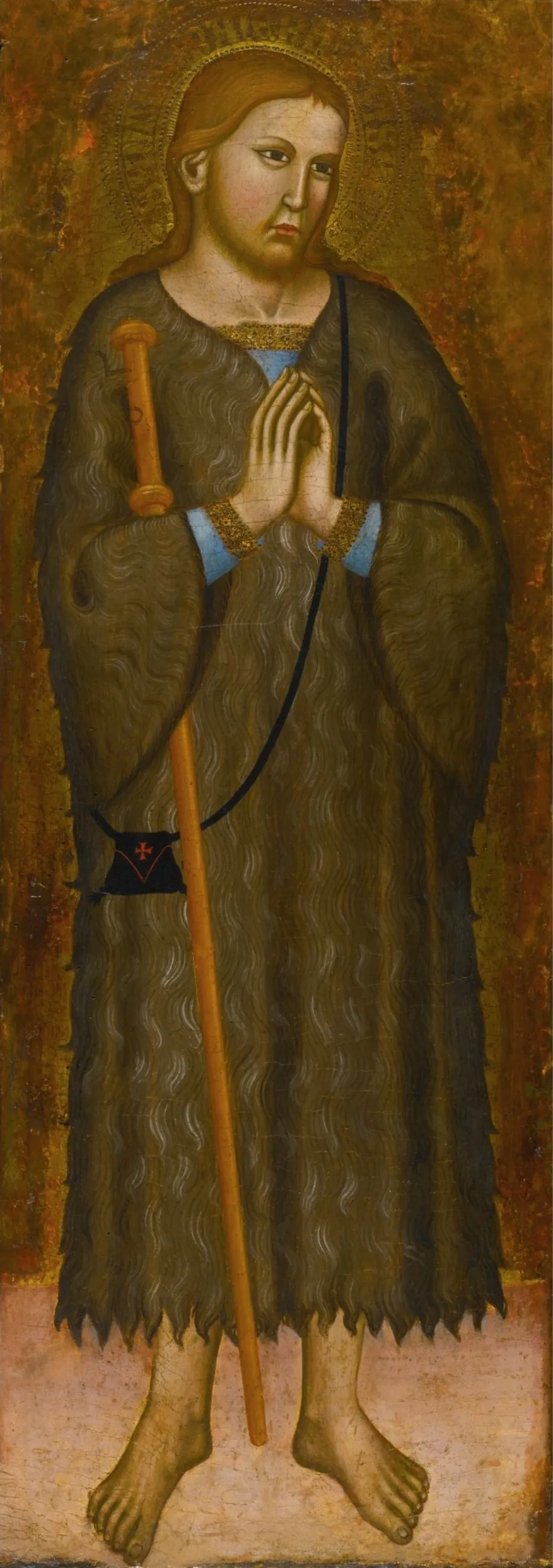 Italian Gothic panel painting on a gold ground of a young man with long reddish hair and a halo, barefoot, hands joined in prayer, wearing a shaggy brown hair tunic to his shins, with a tall staff and a small black bag marked with a red cross