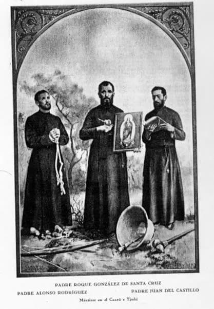 Black-and-white print of three bearded Jesuits in cassocks standing in a landscape under an ornamental arch: the one on the left holds a coiled rope, the one in the center holds up a framed picture of the Virgin Mary, and the one on the right holds an open book, with a fallen bell and weapons at their feet. The caption names Roque González de Santa Cruz, Alonso Rodríguez and Juan del Castillo, martyrs at Caaró and Ijuí, with Roque's name centered above the others