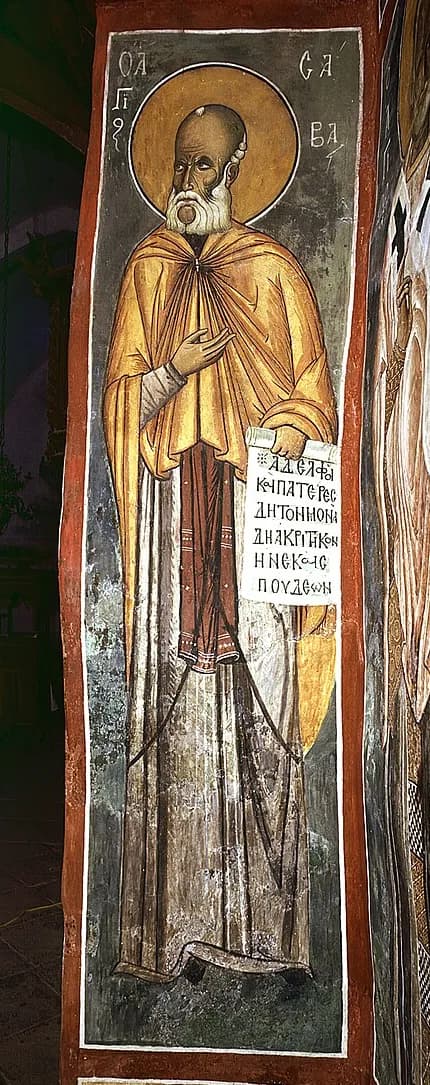 Tall narrow wall painting of an old, bald monk with a short white beard and a gold halo, in an ochre cloak over a pale tunic, one hand raised to his chest and the other holding an open scroll of Greek text, with his name lettered in Greek beside his head